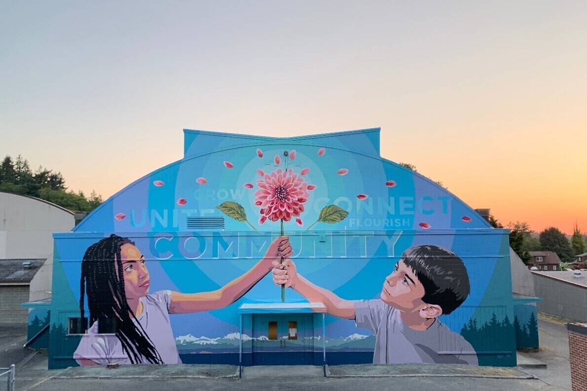 A large mural of two children holding and looking up at a dahlia flower, with a background of blues and lavender circles. It covers the end of a large arched building.