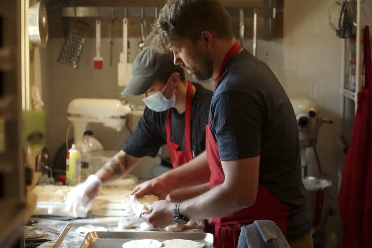 Two adults wear red aprons and prepare take-out meals in kitchen. 